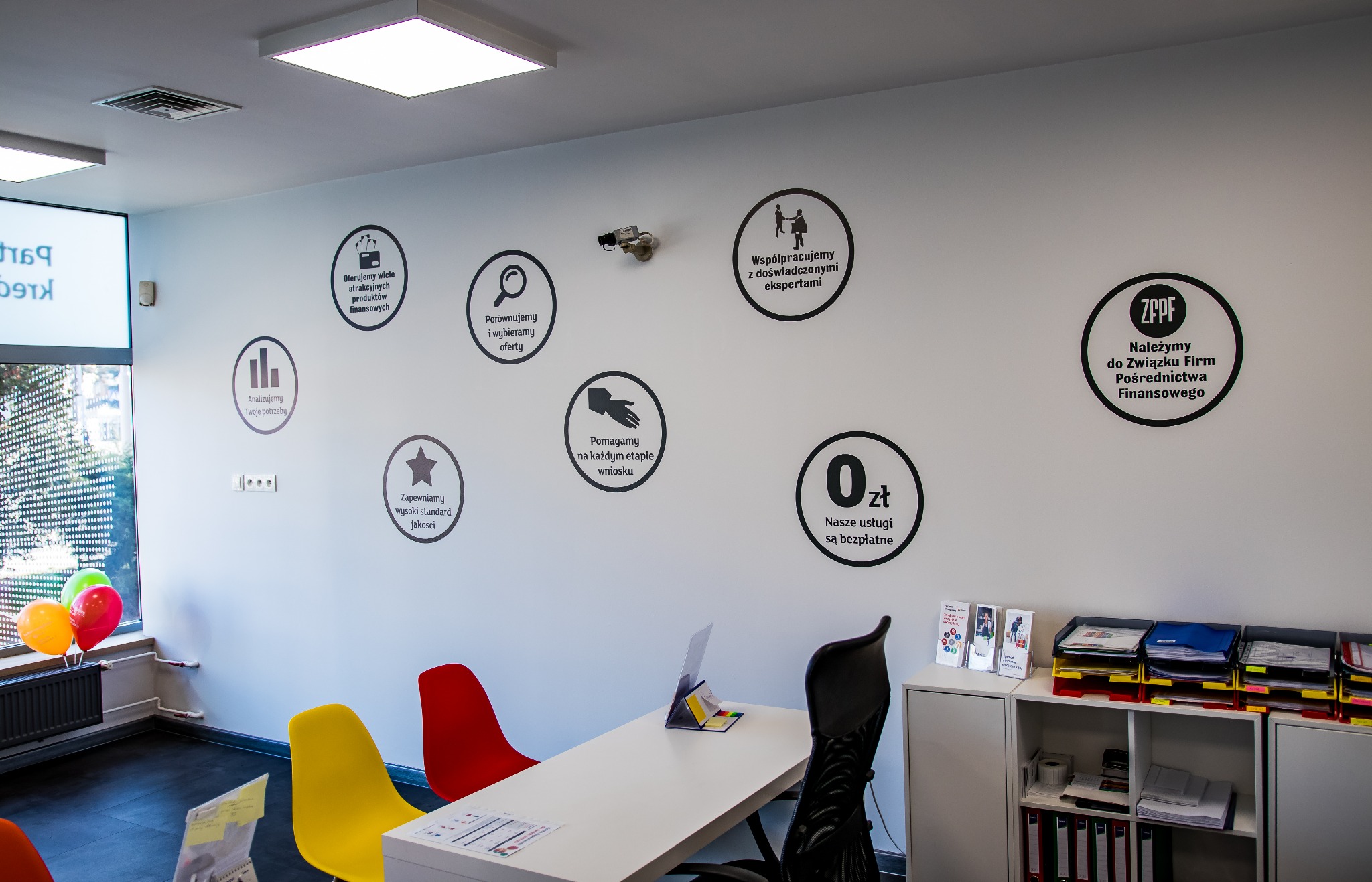 Biała ściana biura z czarnymi okrągłymi naklejkami informacyjnymi o usługach finansowych, pod nimi biurko z kolorowymi krzesłami i regał z dokumentami.