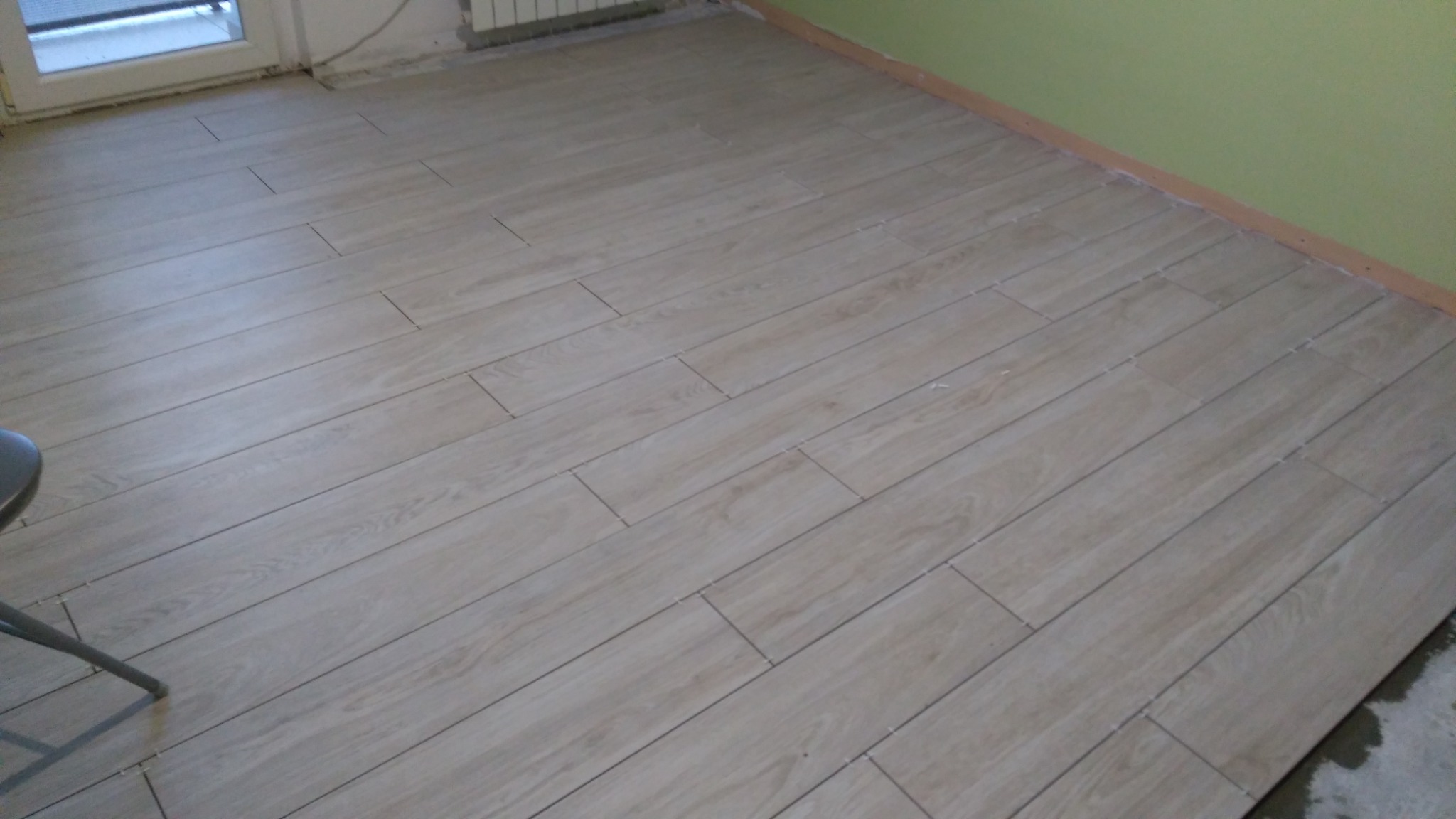 Ułożona podłoga z płytek imitujących drewno, widoczne krzyżyki dystansowe, fragment krzesła w lewym dolnym rogu, zielona ściana w tle.