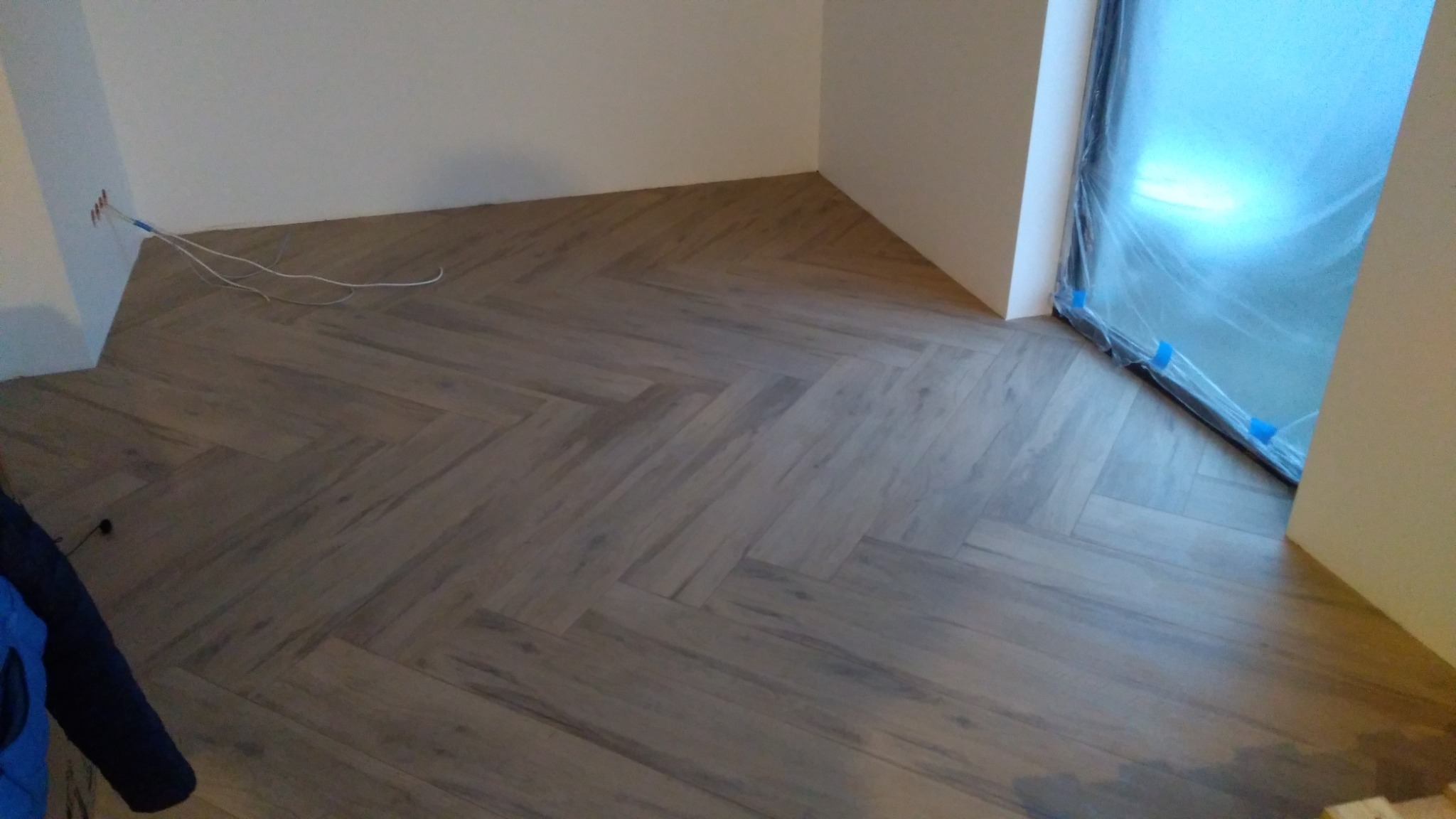 Ułożona pod kątem jodełki parkietowa podłoga w trakcie remontu, okno zabezpieczone folią malarską, wystające przewody elektryczne ze ściany.
