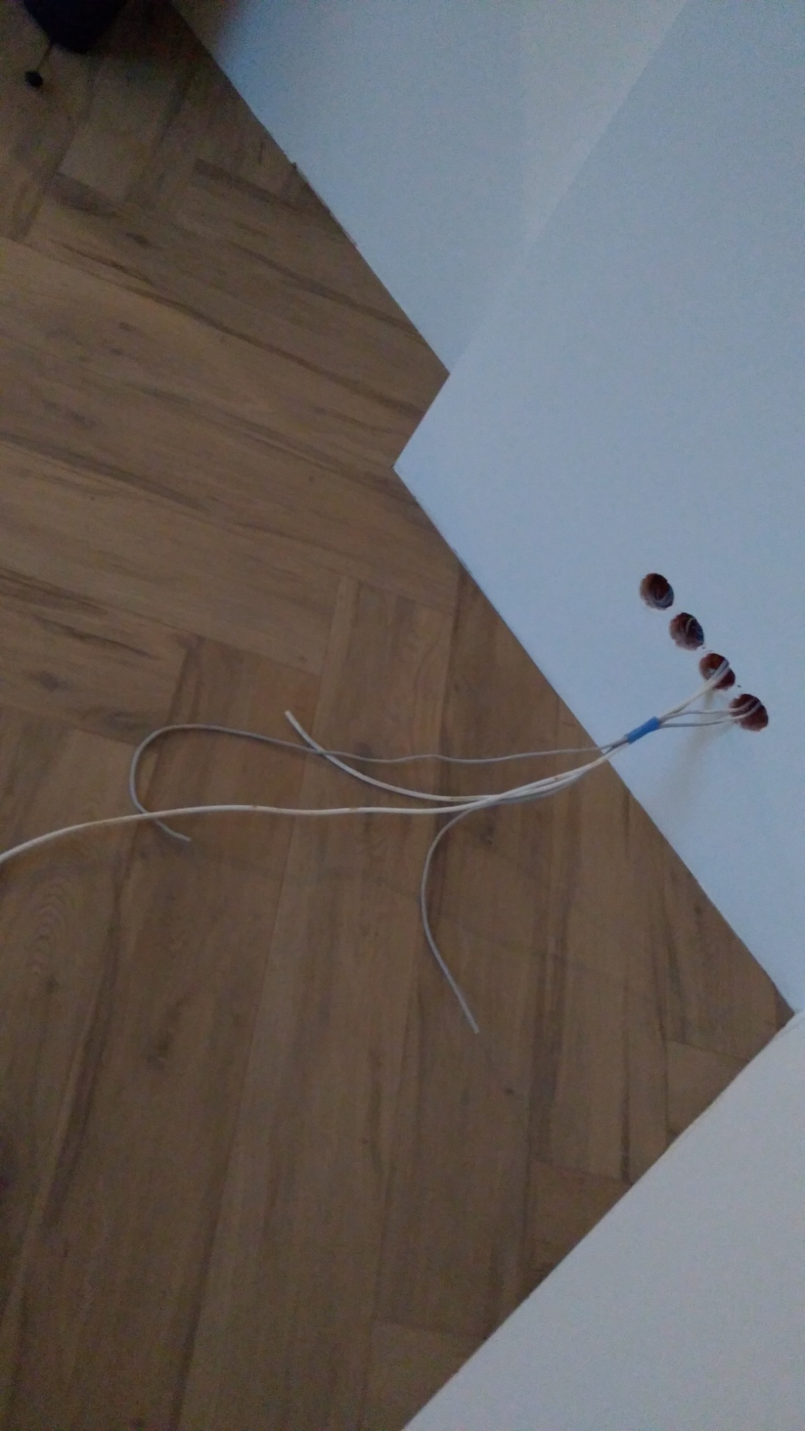 Świeżo wywiercone otwory w ścianie, z których wystają zabezpieczone taśmą przewody elektryczne, ułożone na panelach podłogowych w odcieniach brązu.