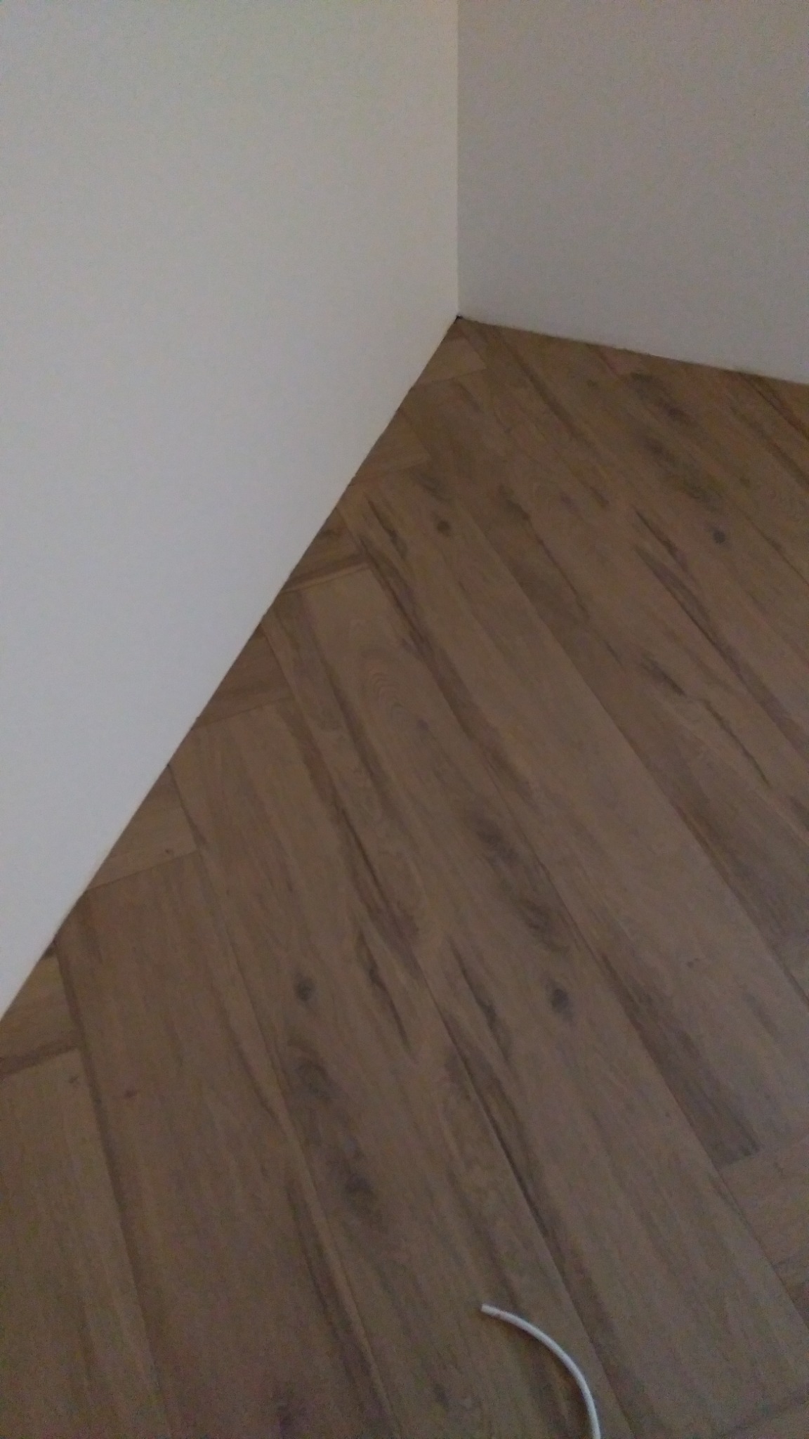 Ukończone połączenie podłogi z paneli w rogu pomieszczenia, widoczny fragment białej ściany i wystający kabel przy listwie przypodłogowej.