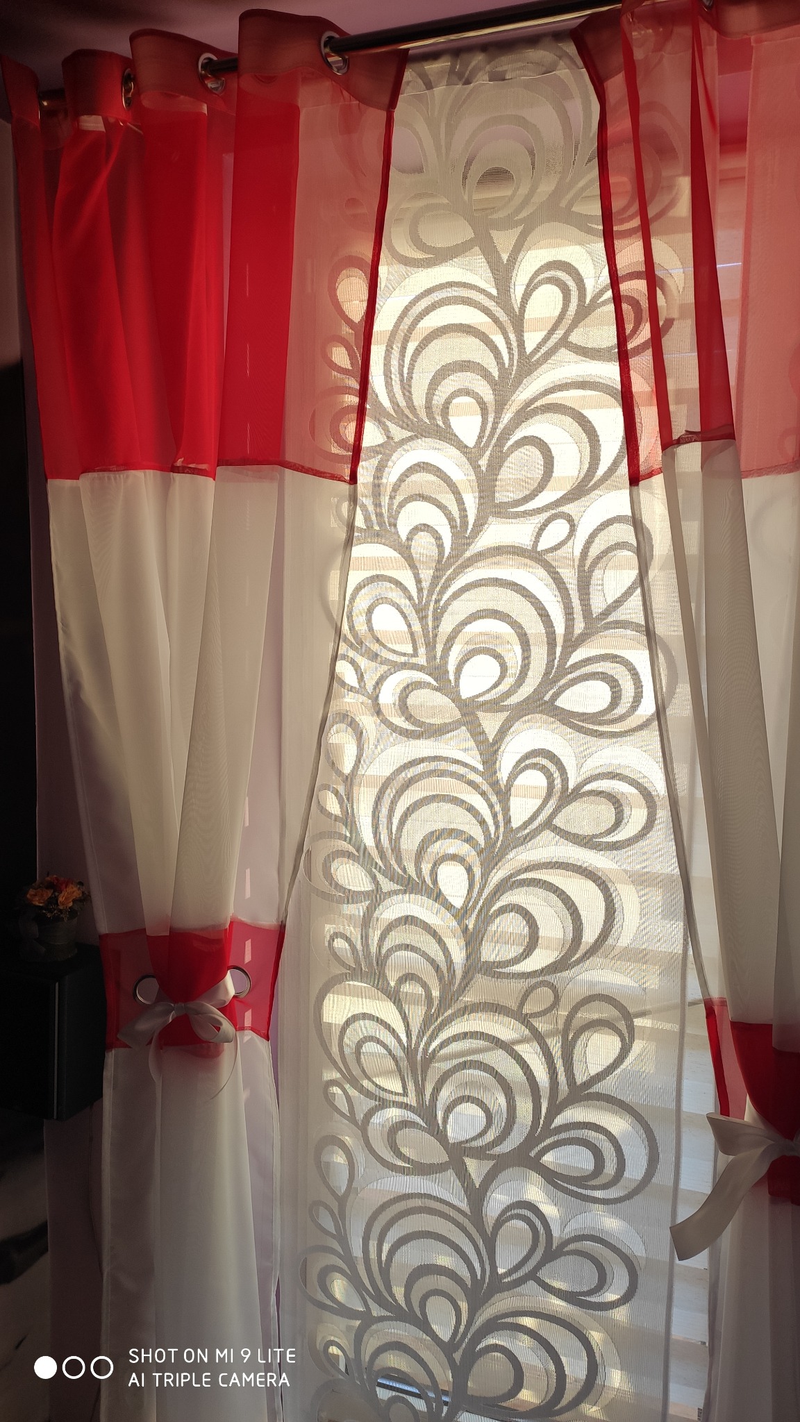 Delikatna, półprzezroczysta firanka z motywem roślinnym w odcieniach szarości, zestawiona z czerwono-białymi zasłonami, upiętymi satynową kokardą.