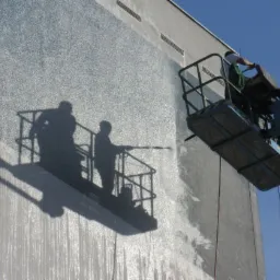 Pracownik na podnośniku koszowym myje elewację budynku za pomocą myjki ciśnieniowej, cień kosza z pracownikami widoczny na elewacji.