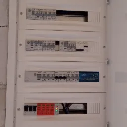 Otwarta skrzynka rozdzielcza z widocznymi bezpiecznikami EATON, Schneider Electric oraz zabezpieczeniami przeciwprzepięciowymi, z żółtą naklejką ostrzegawczą o wyłączeniu zasilania przed zdjęciem...