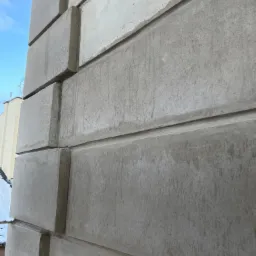 Fragment elewacji budynku z szarych, betonowych bloków o fakturze surowego betonu, widoczne fugi wypełnione zaprawą, niebo w tle.