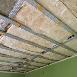 Montaż izolacji z wełny mineralnej w metalowym stelażu sufitu podwieszanego, widoczne żółte taśmy mocujące, ściany pomalowane na zielono.