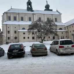 Zimowy krajobraz z barokowym kościołem w tle, pokrytym śniegiem, z trzema zaparkowanymi samochodami na pierwszym planie i drzewem bez liści między nimi.