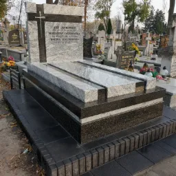 Wielopoziomowy nagrobek z jasnego i ciemnego kamienia Indie Viscont White, z metalowym krzyżem, na cmentarzu z innymi nagrobkami w tle.