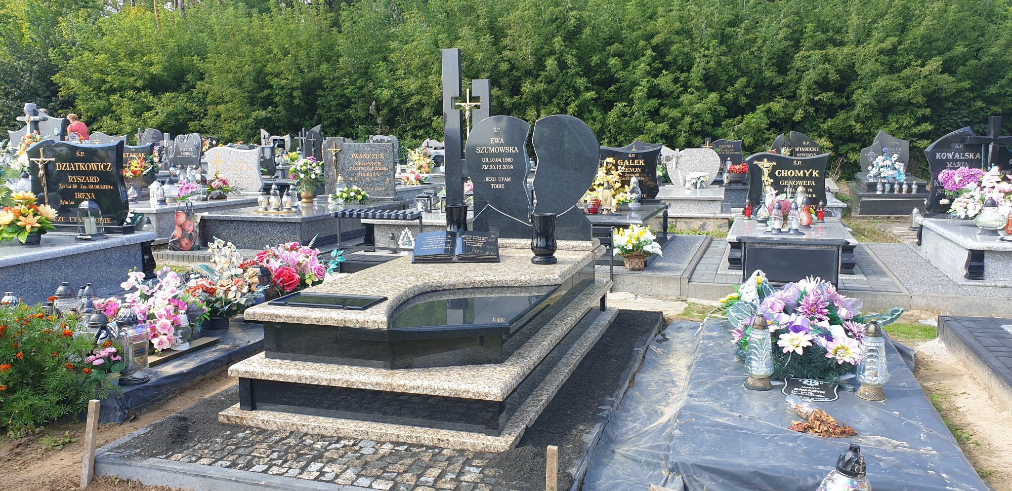 Nowoczesny, dwuosobowy nagrobek z jasnego kamienia z elementami z czarnego granitu na cmentarzu w Tomaszowie Lubelskim. Widoczne otwarte księgi i wazony z ciemnego kamienia. W tle inne groby...