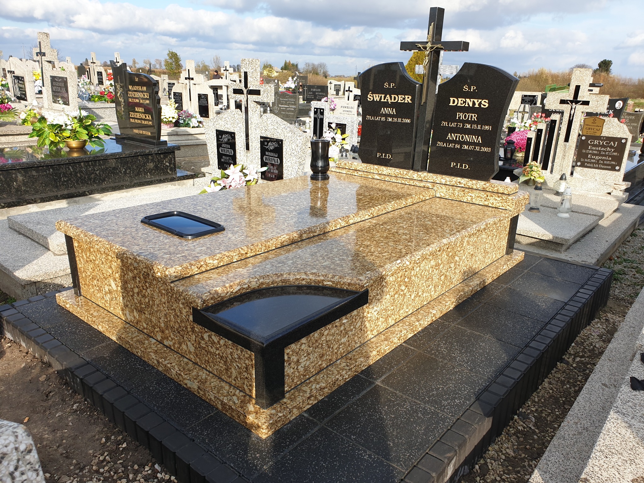 Nagrobek z kamienia Brazylia Giallo Victoria na cmentarzu, z dwoma tablicami imiennymi i czarnym wazonem, otoczony innymi nagrobkami w tle.