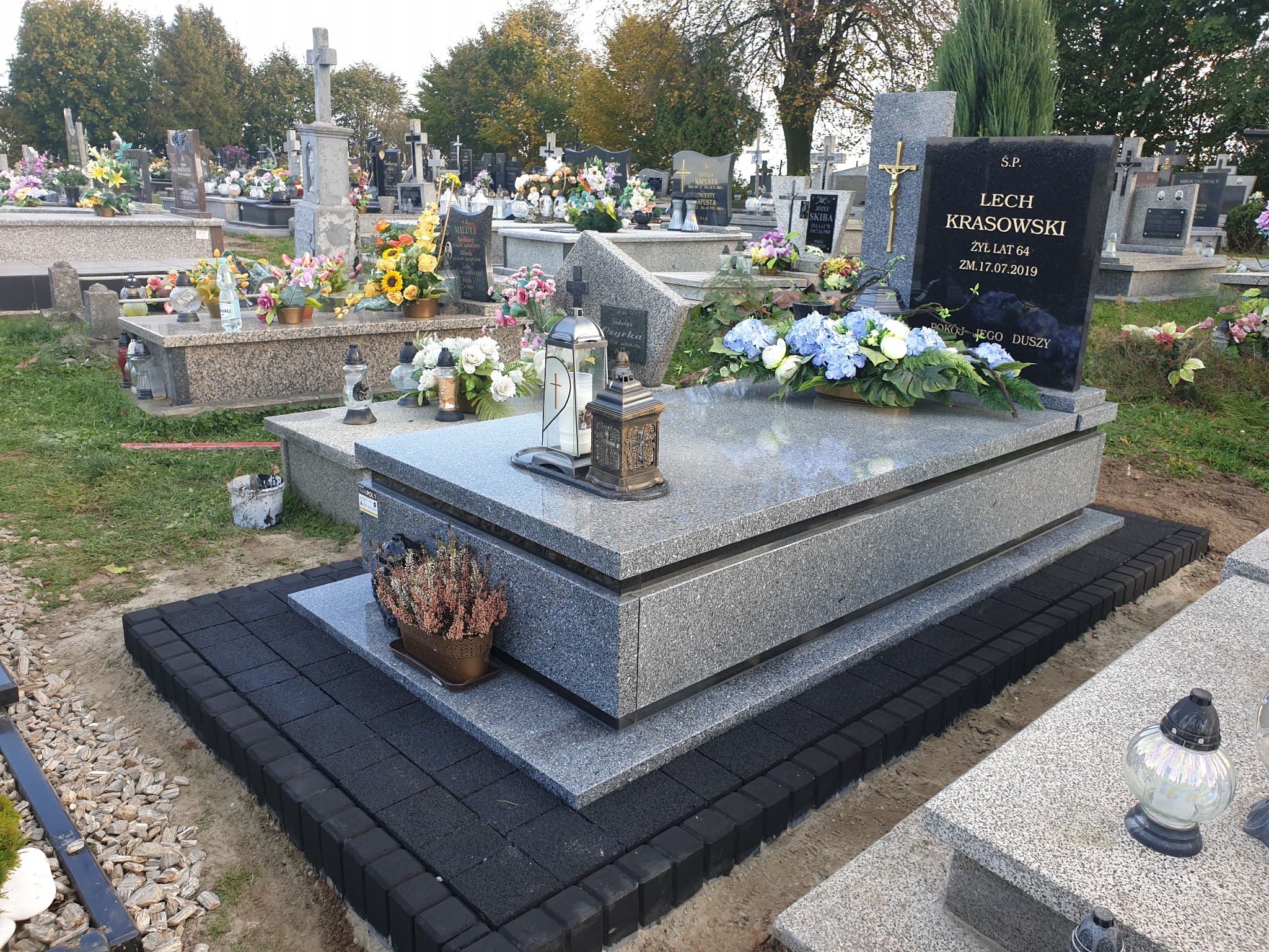 Granitowy nagrobek z czarną tablicą inskrypcyjną, ozdobiony kwiatami i lampionami, otoczony czarną kostką brukową na cmentarzu.