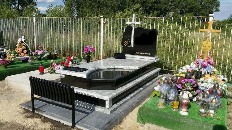 Dwupoziomowy pomnik z jasnego, nakrapianego kamienia z czarnymi elementami, ozdobiony kwiatami i zniczami, z metalową ławeczką obok, na tle metalowego ogrodzenia i zieleni cmentarza.