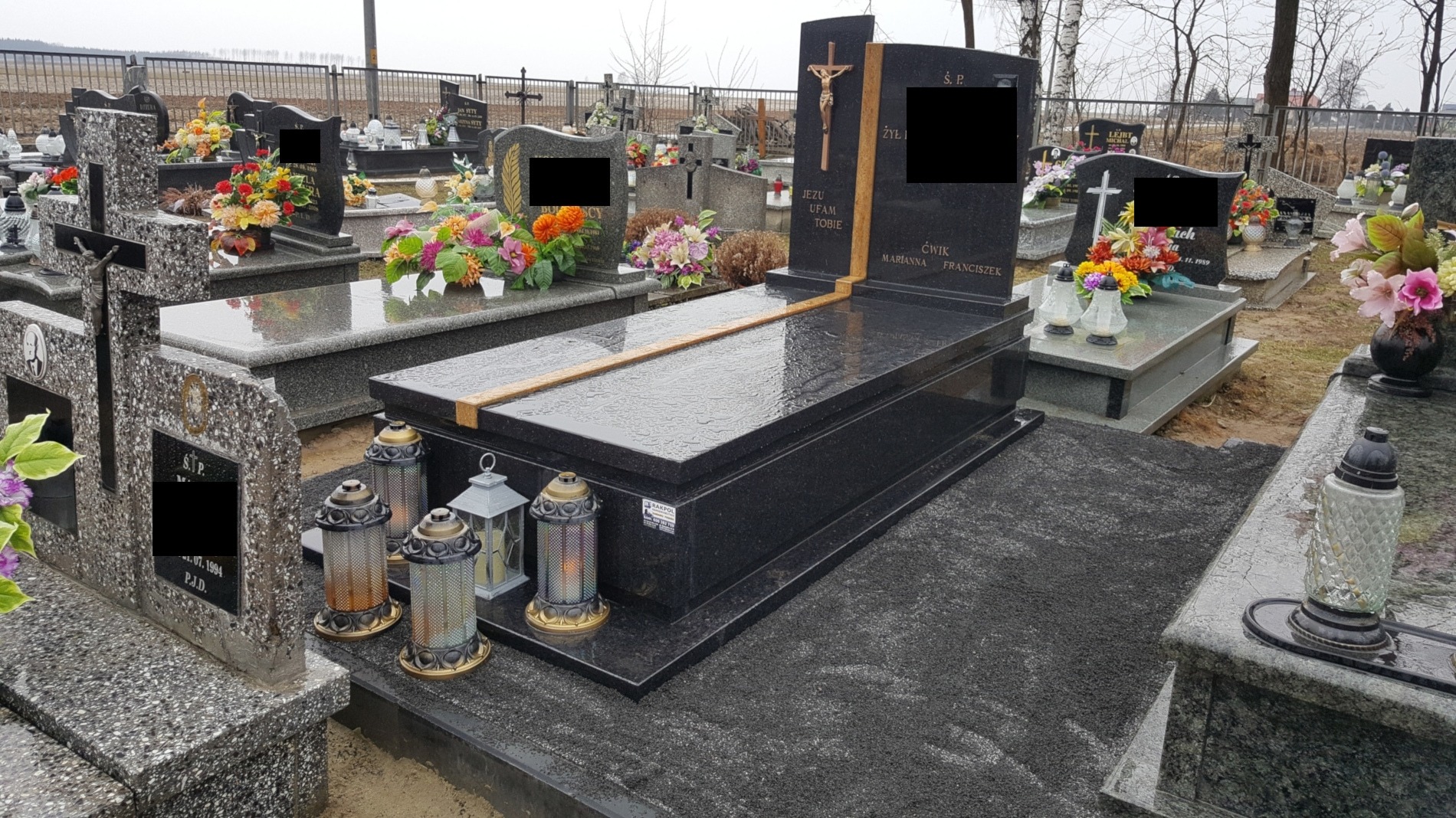 Czarny, polerowany pomnik z szwedzkiego granitu na cmentarzu, z krzyżem i inskrypcjami, otoczony lampionami i kwiatami, widoczny w pochmurny dzień.