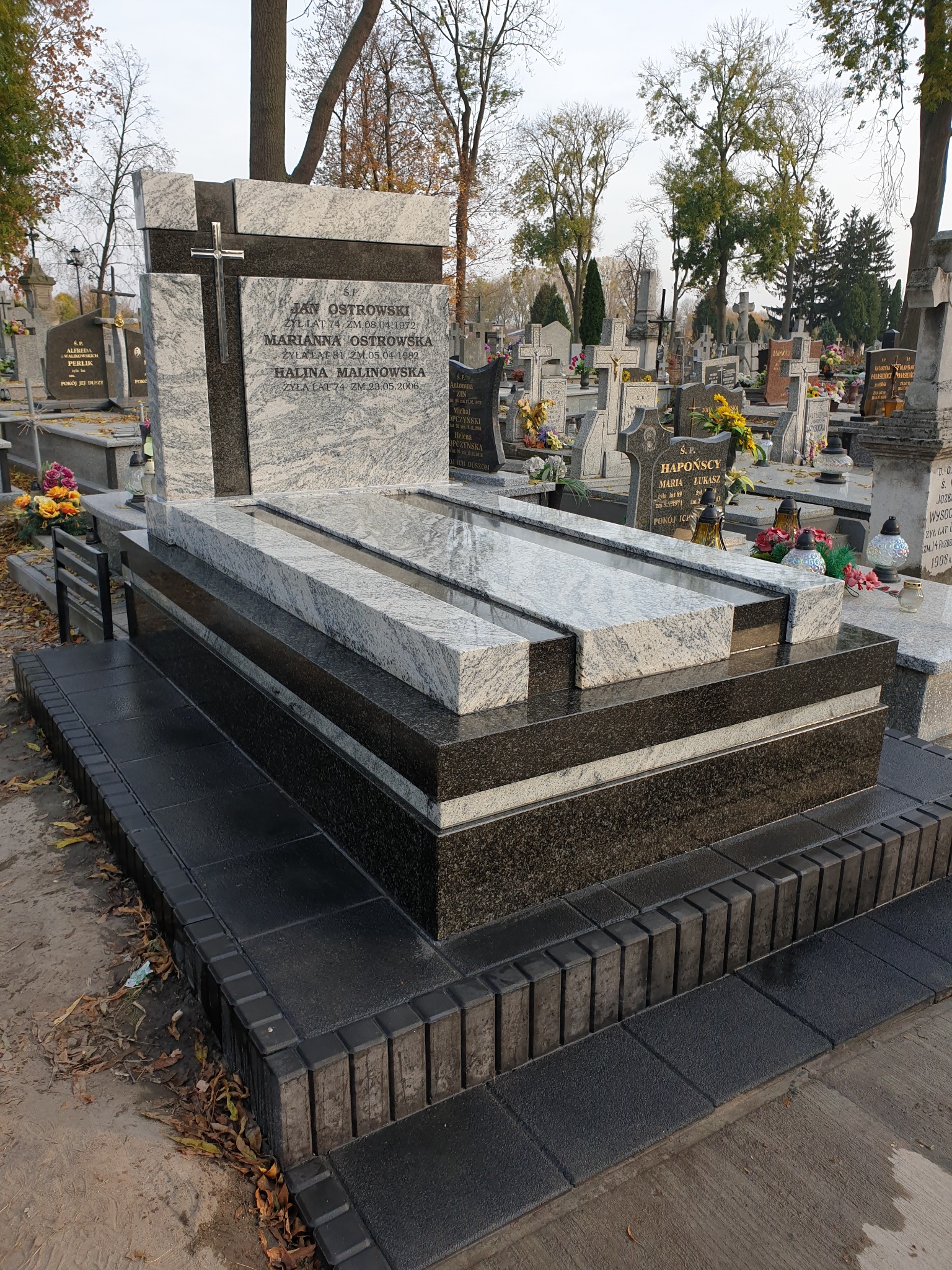 Wielopoziomowy nagrobek z jasnego i ciemnego kamienia Indie Viscont White, z metalowym krzyżem, na cmentarzu z innymi nagrobkami w tle.