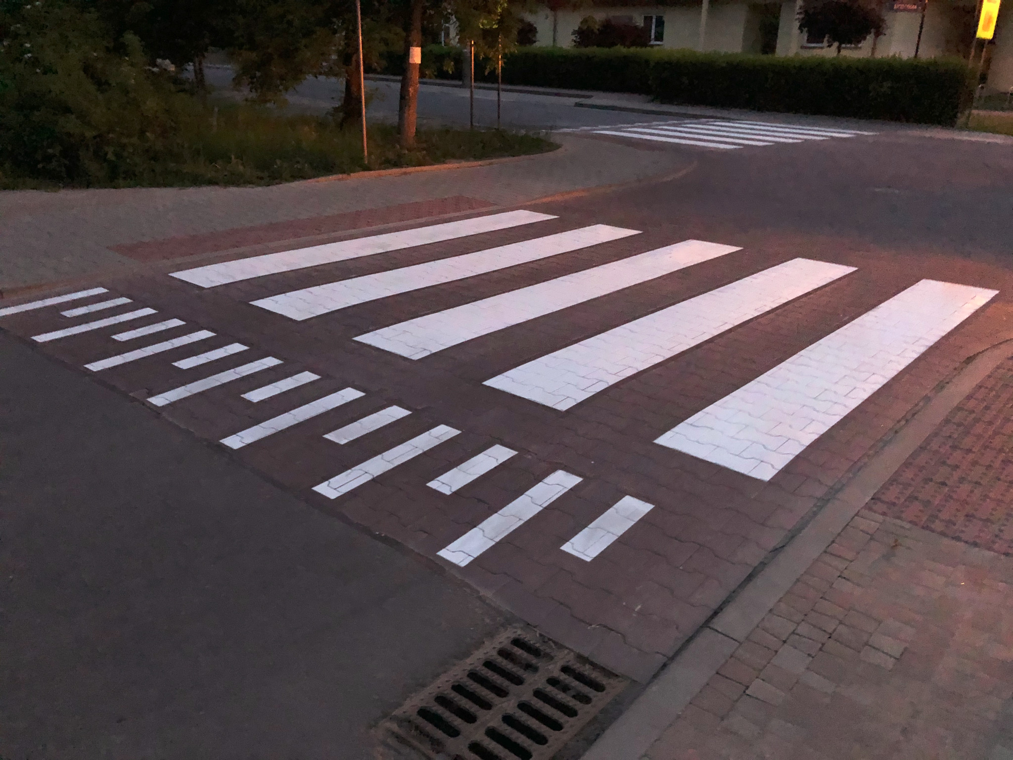 Nowo pomalowane, białe pasy przejścia dla pieszych w Krakowie, z charakterystycznymi, krótkimi liniami po lewej stronie, na tle kostki brukowej i zieleni.