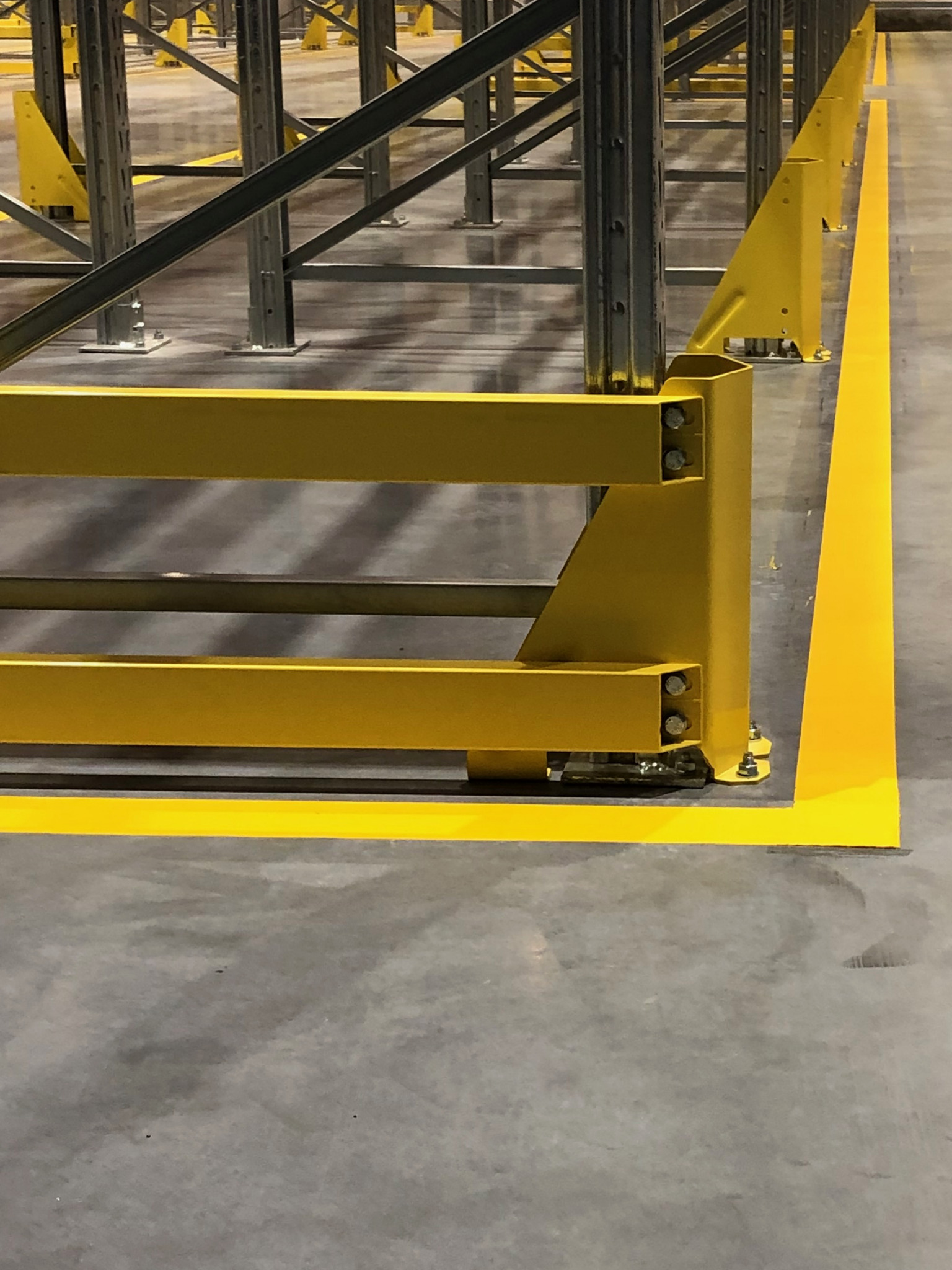 Żółte zabezpieczenia regałów magazynowych i żółte linie na szarej posadzce betonowej.