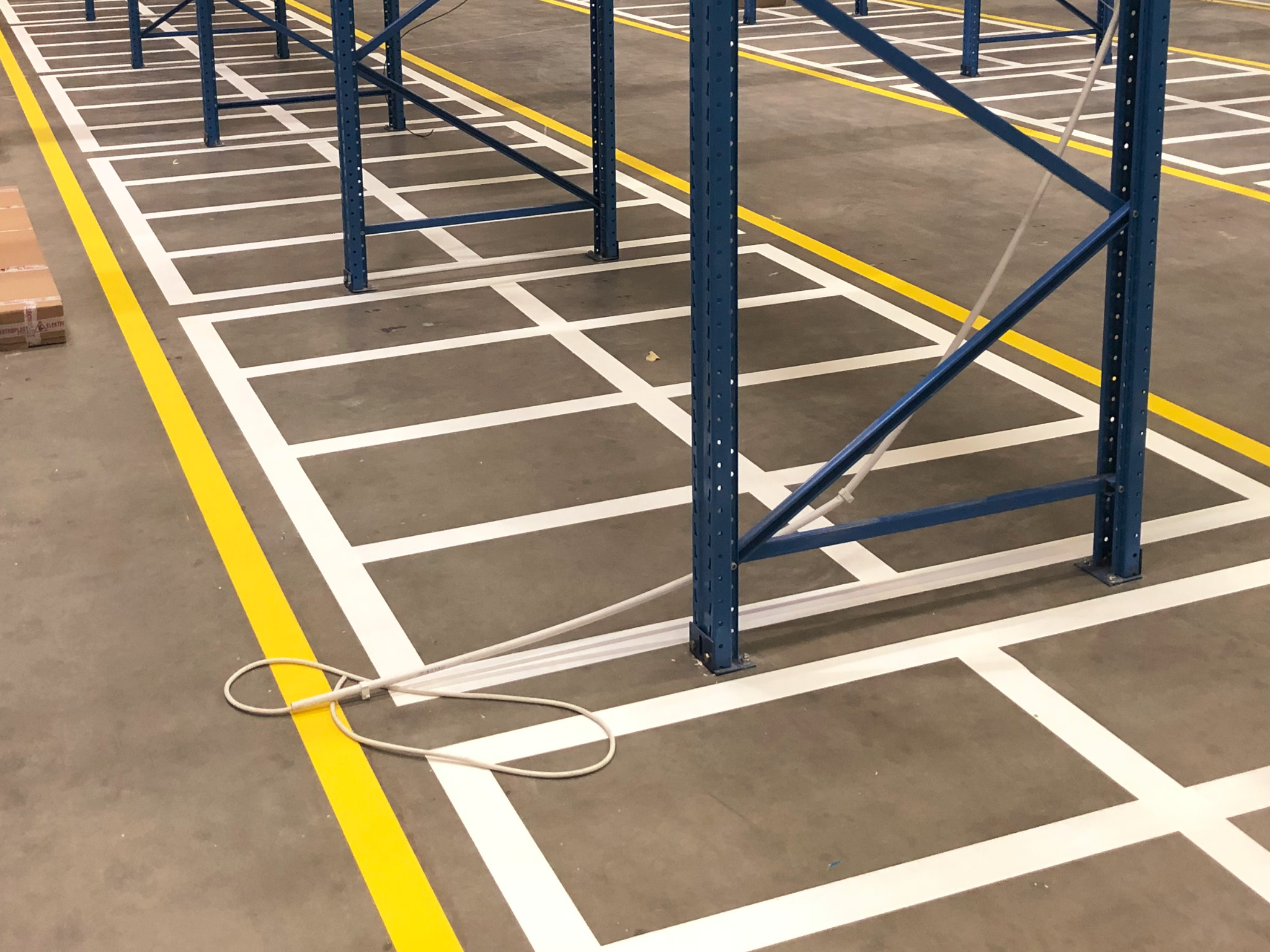 Oznakowanie podłogi w magazynie: białe kwadraty i żółte pasy wyznaczające strefy, widoczne niebieskie elementy regałów, kabel na podłodze.
