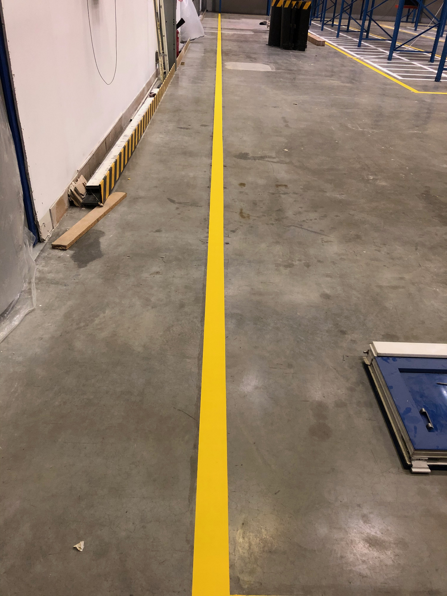 Żółta linia wyznaczająca ciąg komunikacyjny na betonowej posadzce w hali magazynowej, widoczne regały magazynowe w tle.