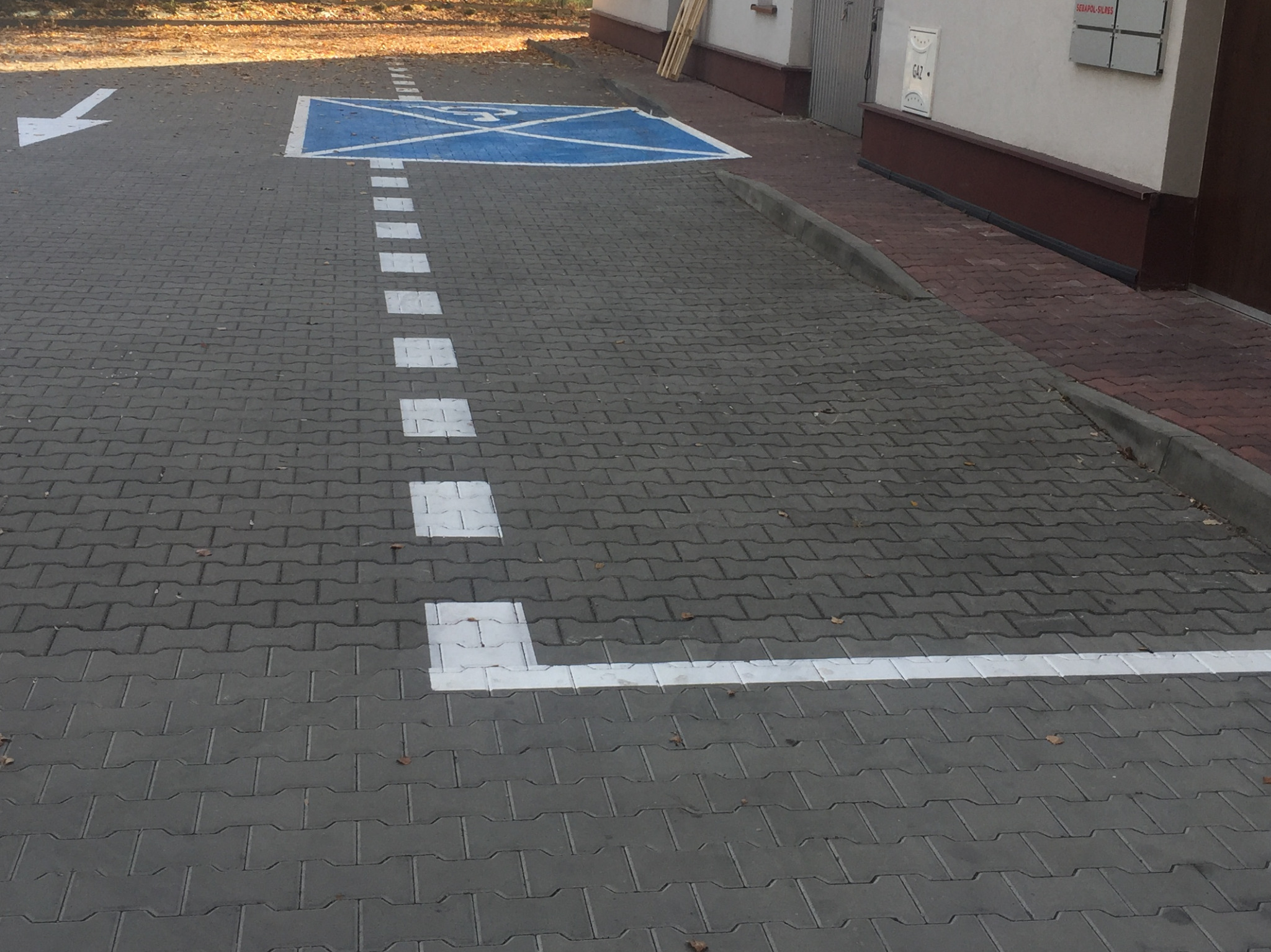 Oznakowane miejsce parkingowe dla osoby niepełnosprawnej, namalowane na szarej kostce brukowej, z widoczną strzałką kierunkową i przerywaną linią oddzielającą stanowiska.