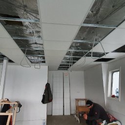 G&K Stroi - Montaż sufitu podwieszanego z płyt kartonowo-gipsowych w pomieszczeniu z dwoma oknami, widoczne przewody i konstrukcja nośna, pracownik siedzi na podłodze