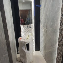 G&K Stroi - Wnętrze łazienki w trakcie prac wykończeniowych: ściany z szarych płytek imitujących marmur, fragment mozaiki, umywalka w folii ochronnej, widoczne przewody elektryczne.