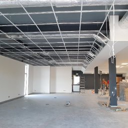 G&K Stroi - Przestronne wnętrze biurowe w trakcie prac wykończeniowych, widoczna stalowa konstrukcja sufitu podwieszanego, instalacja wentylacyjna i pracownik w oddali.