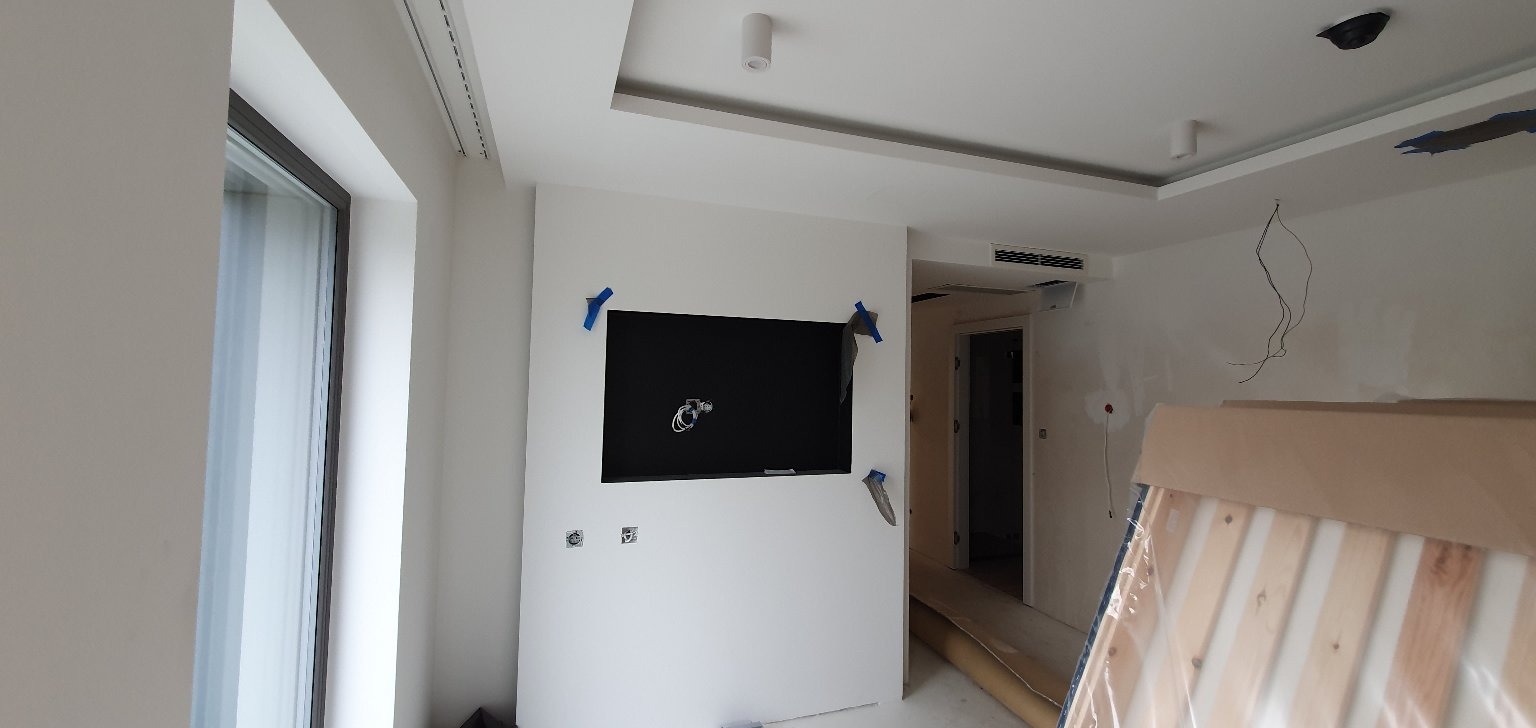 Wnętrze w trakcie prac wykończeniowych: zamontowany telewizor na ścianie, widoczne kable, instalacja elektryczna, elementy sufitu podwieszanego i częściowo zakryte meble w folii ochronnej.