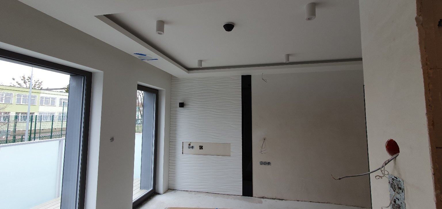 Nowoczesne wnętrze w trakcie wykańczania: biała ściana z dekoracyjną fakturą falistą, wnęka na multimedia, widoczne instalacje elektryczne, okna z widokiem na zewnątrz, sufit podwieszany...