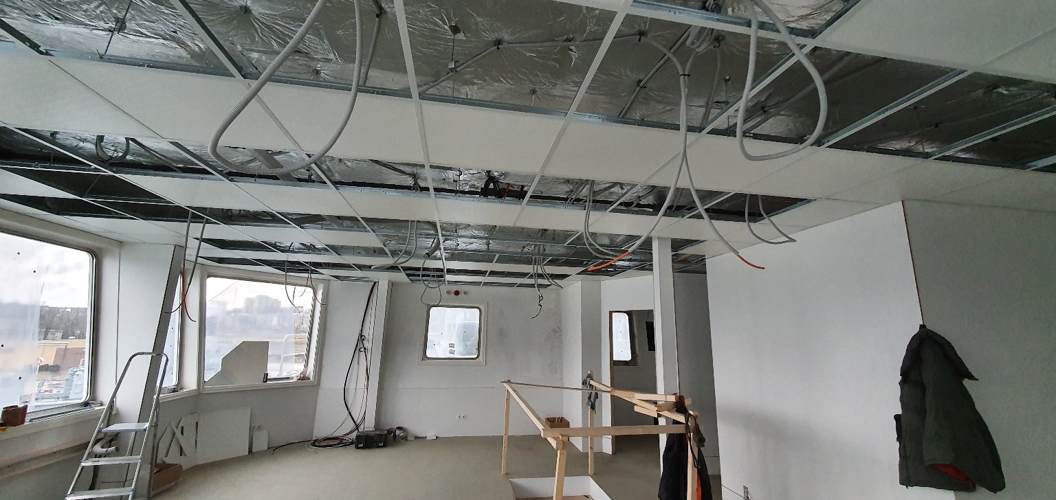 Wnętrze biura w trakcie remontu: widoczne metalowe profile sufitowe, częściowo zamontowane płyty sufitowe, wystające przewody elektryczne, drabina i okna z widokiem na miasto.