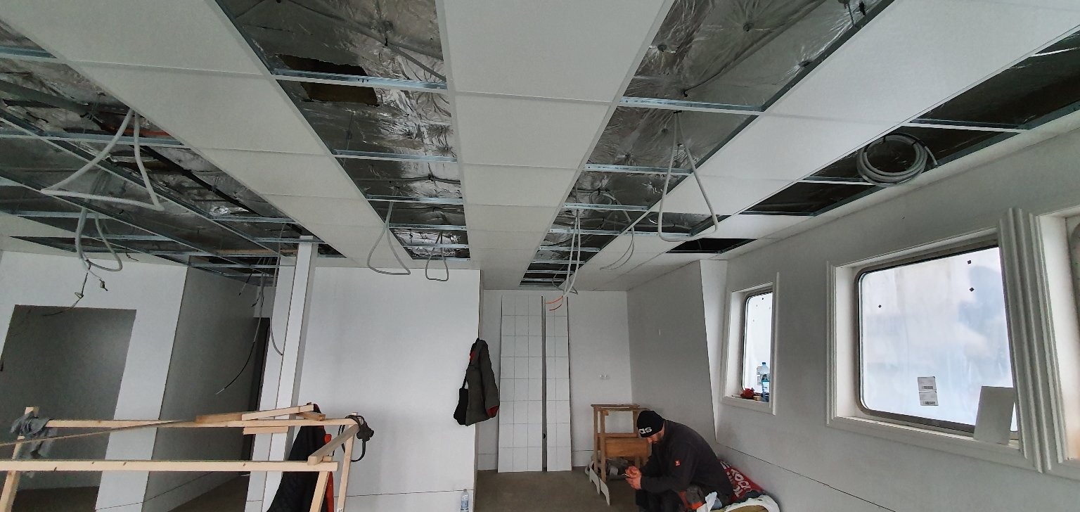 Montaż sufitu podwieszanego z płyt kartonowo-gipsowych w pomieszczeniu z dwoma oknami, widoczne przewody i konstrukcja nośna, pracownik siedzi na podłodze