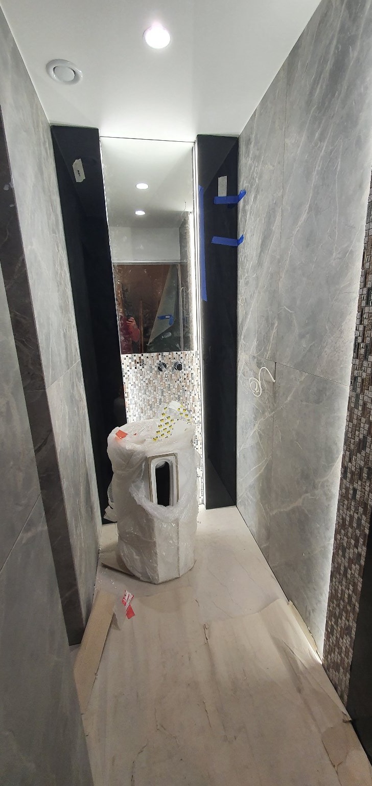 Wnętrze łazienki w trakcie prac wykończeniowych: ściany z szarych płytek imitujących marmur, fragment mozaiki, umywalka w folii ochronnej, widoczne przewody elektryczne.