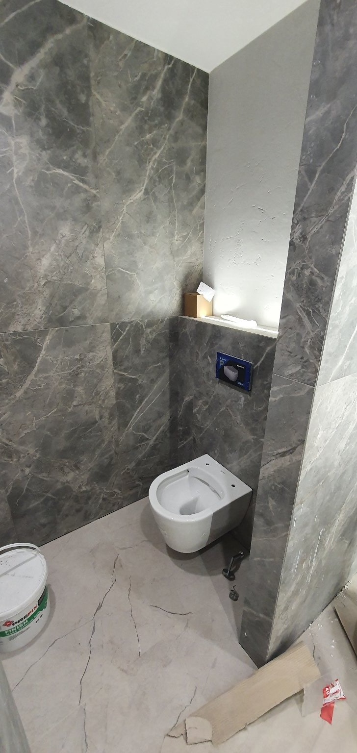 Montaż podwieszanej miski WC na tle szarej ściany imitującej marmur, widoczny pojemnik z masą szpachlową i fragment listwy przypodłogowej.