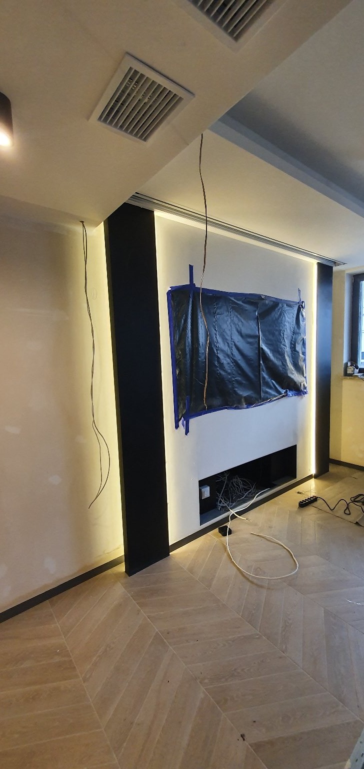 Wnętrze w trakcie modernizacji: ściana z podświetleniem LED, zakryta folią ochronną, widoczne przewody, parkiet w jodełkę.