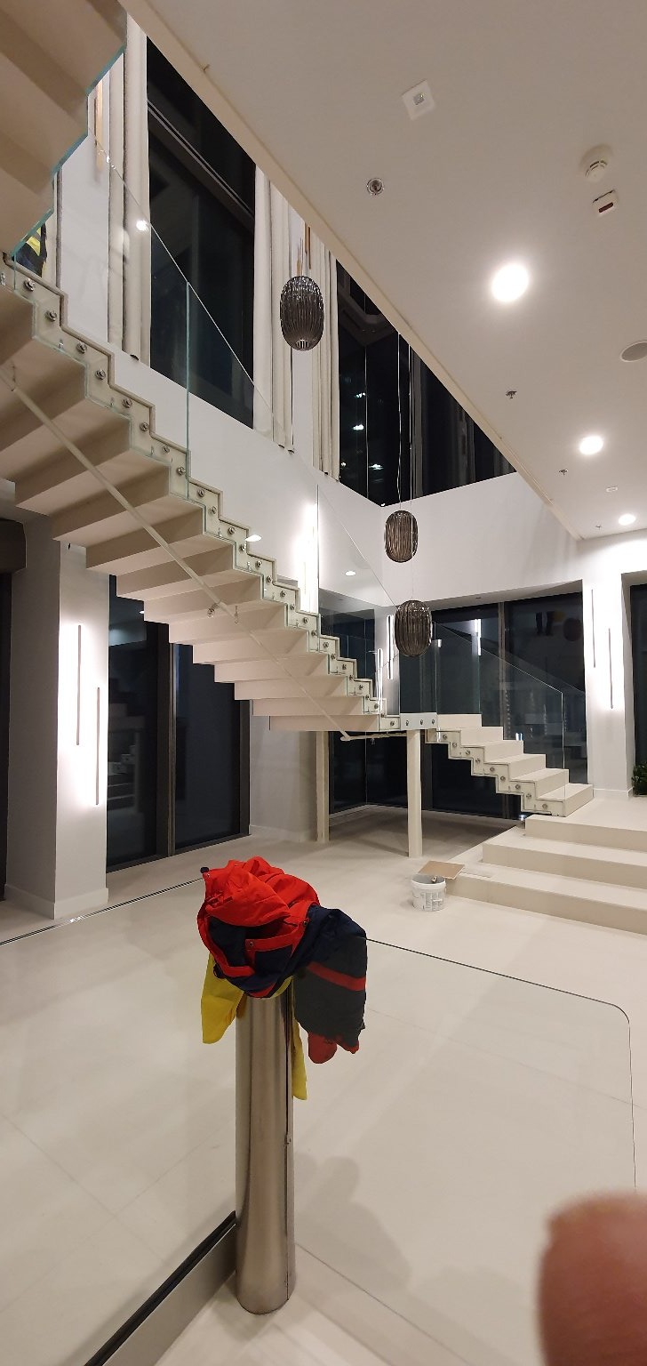 Nowoczesne, białe schody z szklanymi balustradami w przestronnym wnętrzu, wiszące lampy kuliste, odzież robocza na stojaku w tle, widoczne ślady po remoncie.
