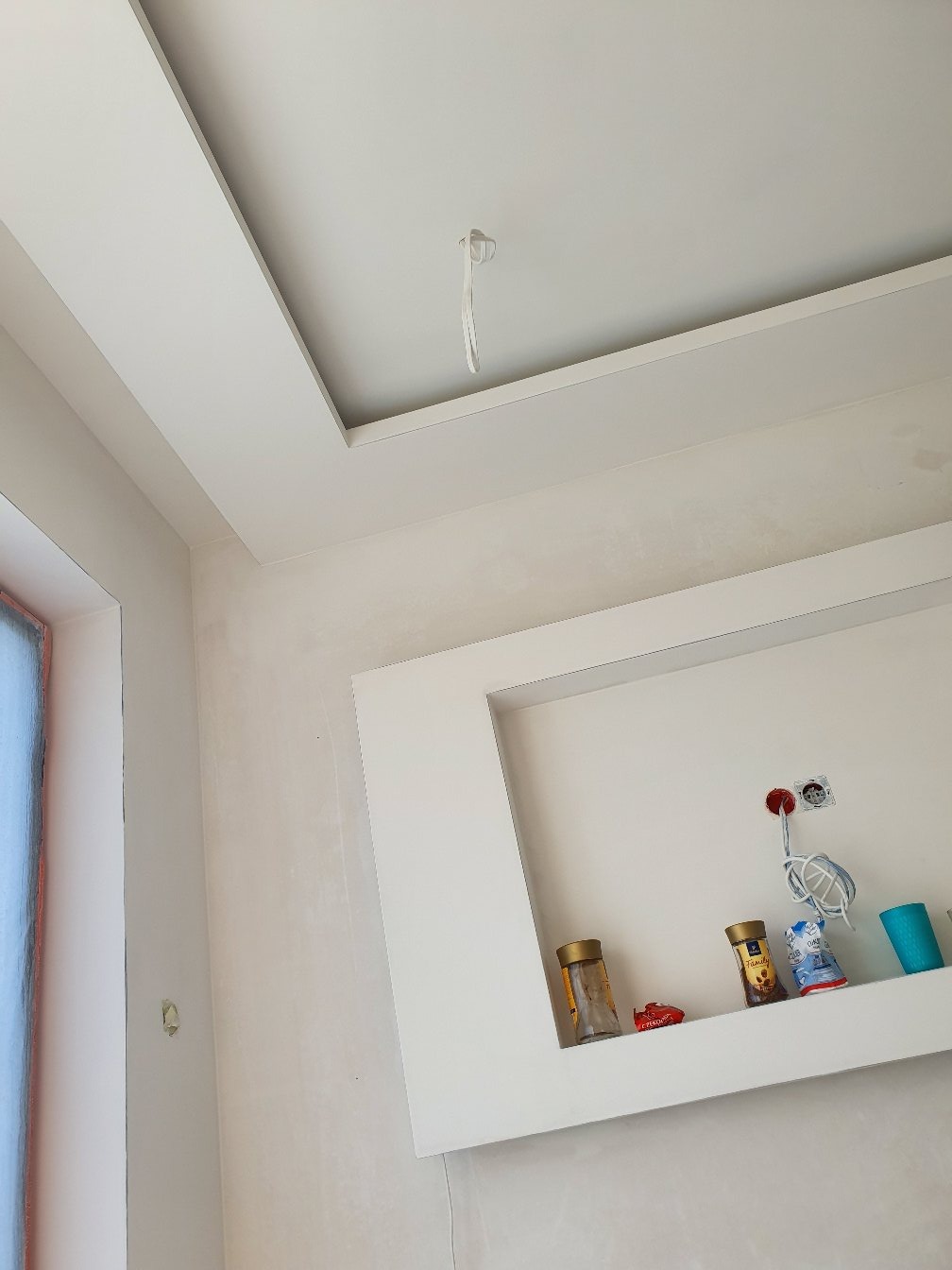 Wnętrze w trakcie prac wykończeniowych: widoczny sufit podwieszany z wnęką, ściany przygotowane do malowania i wnęka na ścianie z instalacją elektryczną i półką z drobnymi przedmiotami.