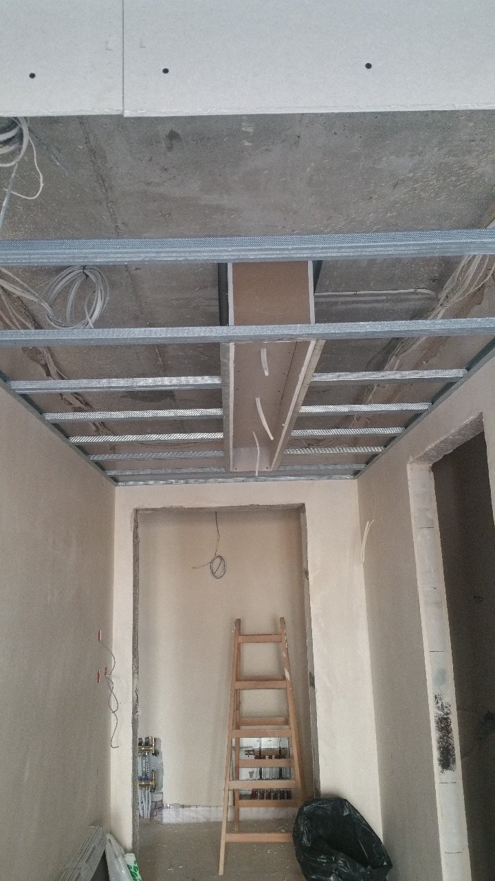 Montaż metalowej konstrukcji sufitu podwieszanego z widocznymi instalacjami elektrycznymi, drabina oparta o ścianę w surowym stanie.