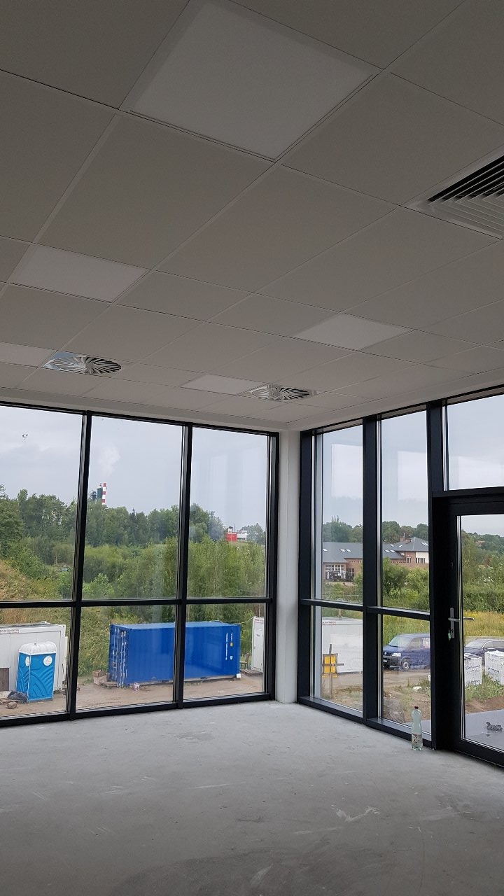 Wnętrze biura z nową podwieszaną kasetonową sufitą, widok na zewnątrz przez duże okna z czarnymi ramami, na zewnątrz widoczne kontenery i zieleń.