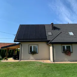 Dom jednorodzinny z panelami fotowoltaicznymi na części dachu, widok z ogrodu w słoneczny dzień.