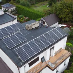 Widok z góry na instalację paneli słonecznych na dachu domu jednorodzinnego, obok leżą kartony, a obok nich stoi osoba w szarym ubraniu, dodatkowo na dachu innego budynku również widać panele...