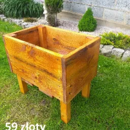Drewniana skrzynia na rośliny, lakierowana na złoty kolor, ustawiona na trawniku przed murem z betonowych bloczków. Widoczny napis '59 złoty'.