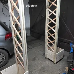 Niedokończona drewniana konstrukcja przypominająca pergolę z kratownicą i donicami u podstawy, częściowo pomalowana na biało, widoczna w garażu z samochodem w tle i napisem '360 złoty' na górze.