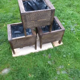 Trzy poziomy, brązowe, drewniane donice ogrodowe z czarnym wkładem, ustawione na trawie, z widoczną ceną '42 złote' na górze zdjęcia.