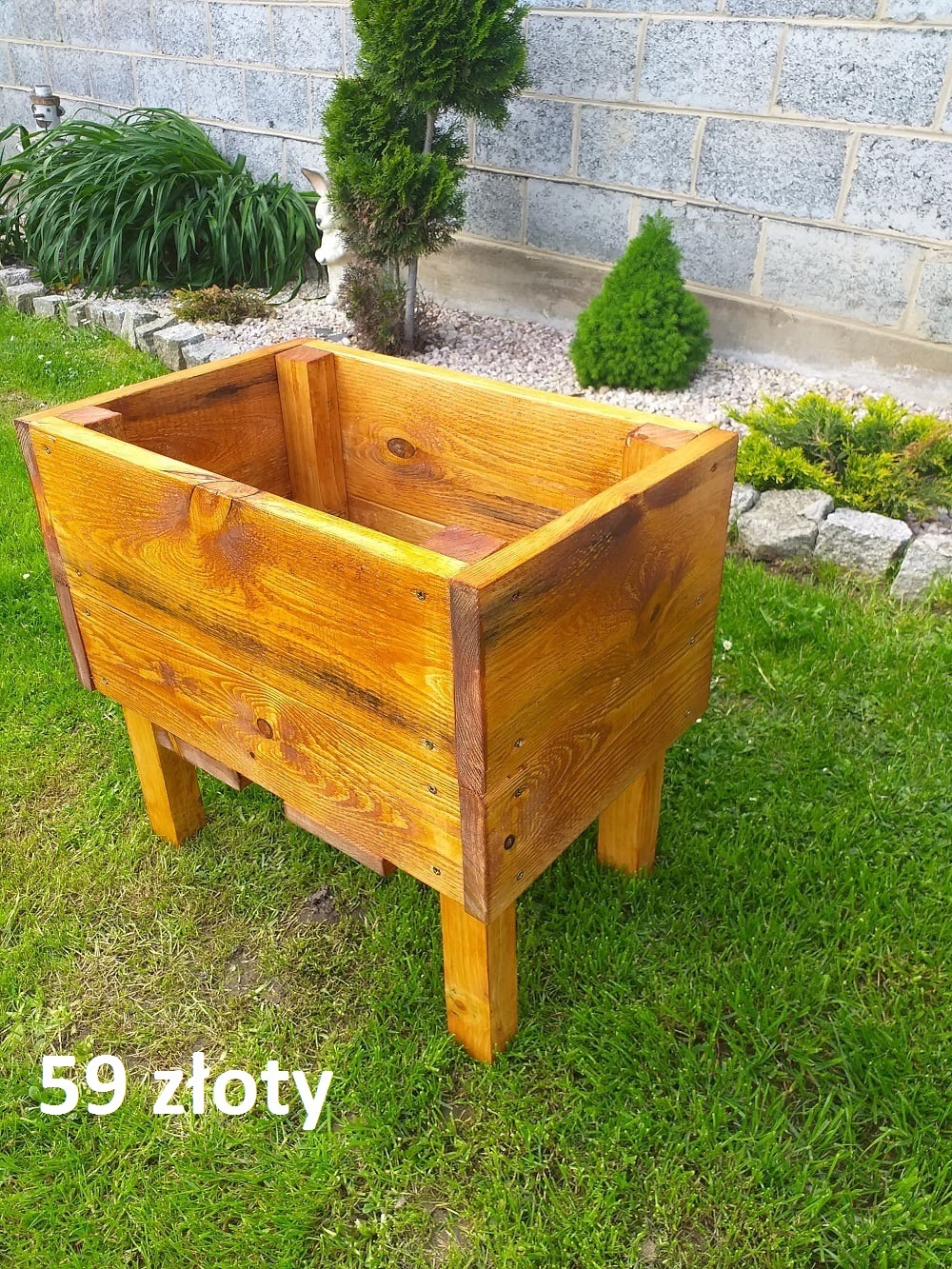 Drewniana skrzynia na rośliny, lakierowana na złoty kolor, ustawiona na trawniku przed murem z betonowych bloczków. Widoczny napis '59 złoty'.