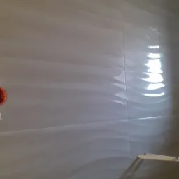 Ściana wyłożona błyszczącymi, białymi płytkami ceramicznymi z delikatnym, falistym wzorem, widoczny fragment rury i element instalacji.