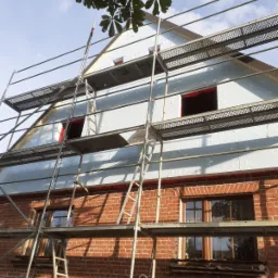 Dom z czerwonej cegły w trakcie ocieplania, widoczne rusztowania i warstwa izolacyjna na poddaszu, częściowo zakryta przez liście drzewa.