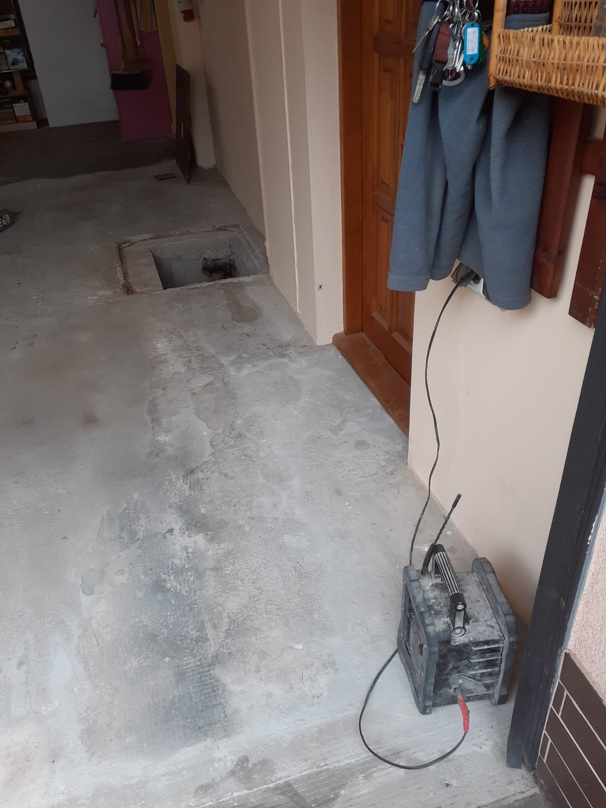 Surowy betonowy podkład podłogowy w pomieszczeniu gospodarczym z otwartym otworem rewizyjnym, widoczny sprzęt elektryczny podłączony do gniazdka.