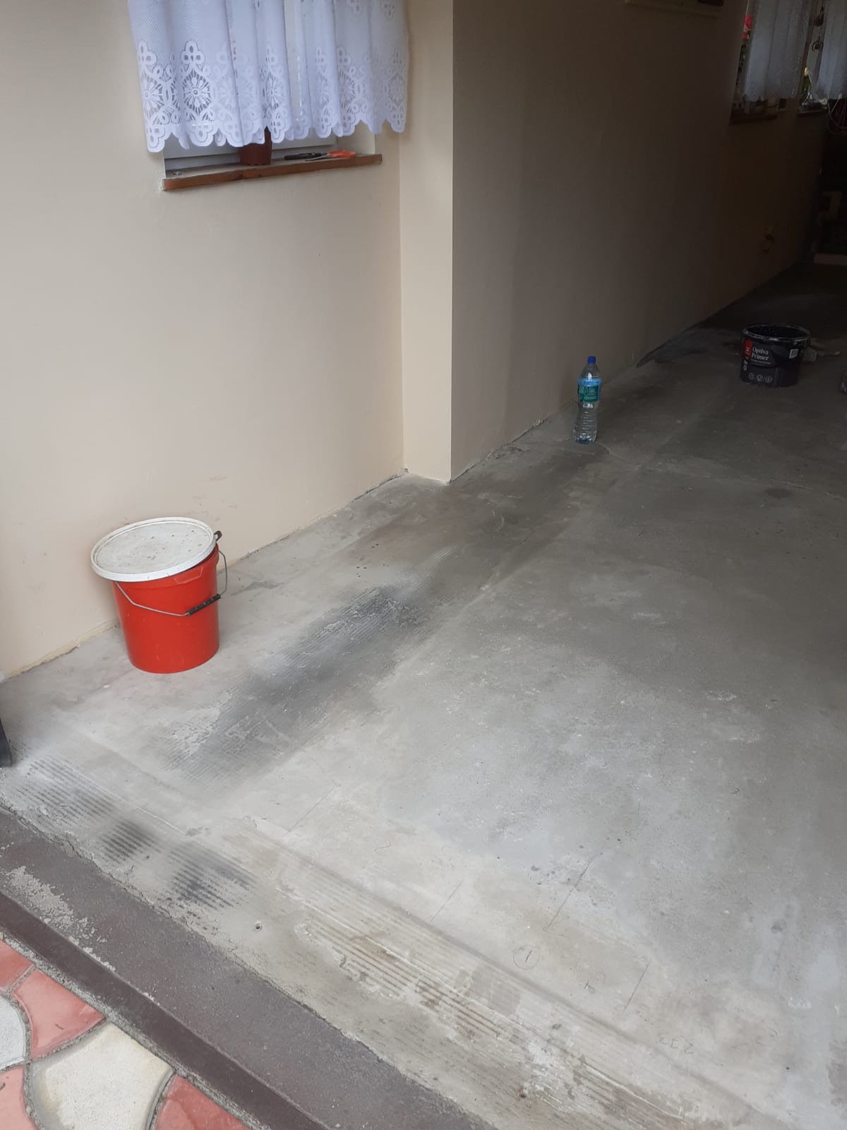 Pomieszczenie gospodarcze w trakcie przygotowania do prac wykończeniowych: gruntowanie ścian w beżowym odcieniu, widoczny czerwony kubeł z pokrywą, butelka wody mineralnej i wiadro z gruntem...