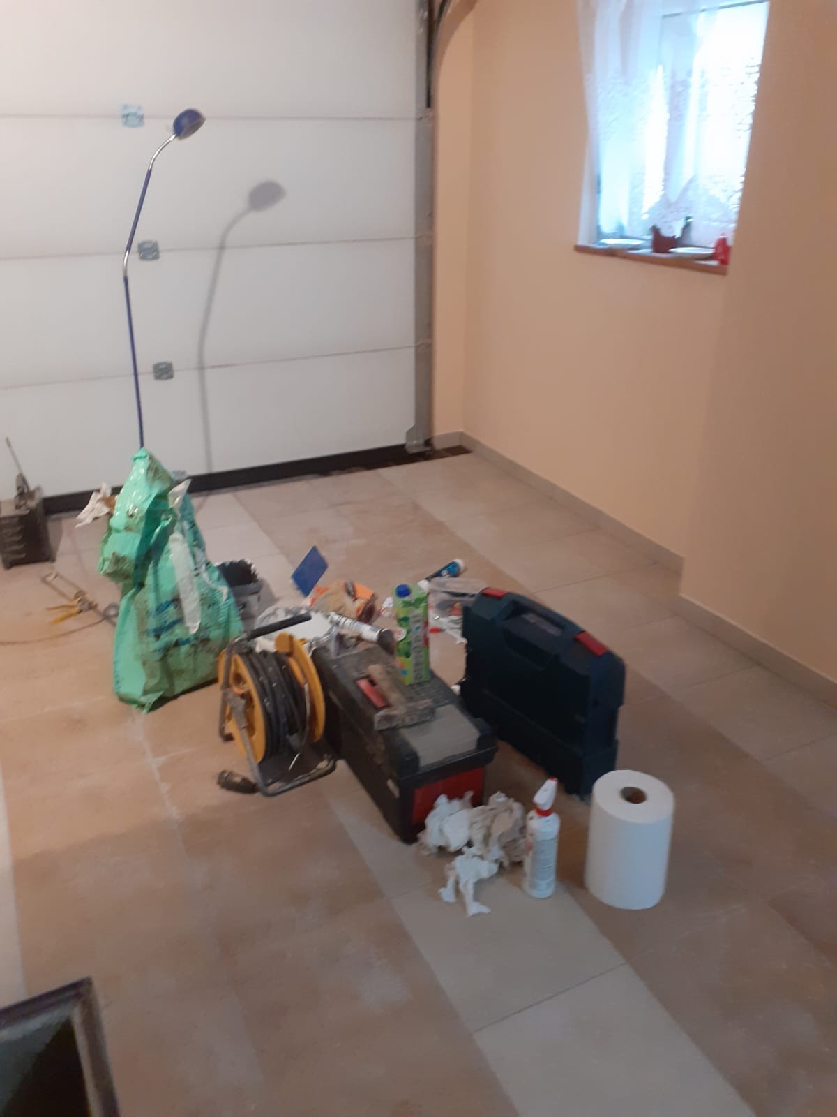 Pomieszczenie gospodarcze w trakcie prac wykończeniowych: widoczna brama garażowa, okno z firanką, beżowe ściany, płytki na podłodze, torba z zaprawą, skrzynka narzędziowa, przedłużacz na bębnie...