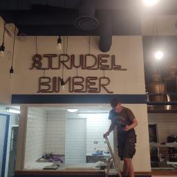 Nla Digital Group sp. z o.o. - Pracownik na drabinie, prawdopodobnie podczas wykańczania wnętrza lokalu gastronomicznego z białą cegłą, widoczne napisy 'Strudel Bimber' wykonane w formie instalacji z rurek, surowe żarówki...