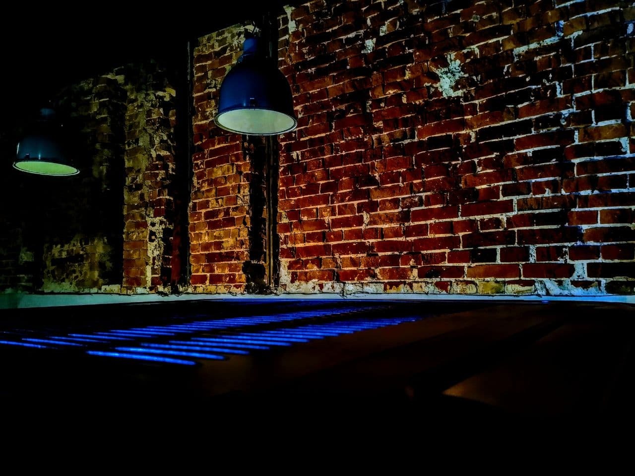 Ciemne wnętrze z odsłoniętą, czerwoną cegłą na ścianie, oświetlone wiszącymi lampami przemysłowymi, z niebieskimi refleksami światła na podłodze.